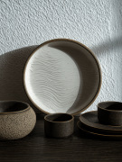Duży ceramiczny talerz z pofalowanym, strukturalnym wzorem przypominającym piasek opiera się o białą, chropowatą ścianę. W mocnym świetle słonecznym otacza go dopasowany zestaw naczyń w ciemnobrązowym kolorze, w tym małe czarki, miska i spodki.