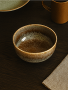 Okrągła ceramiczna miska z zacieranym, brązowo-beżowym szkliwem stoi na ciemnym drewnianym stole. Przytulna aranżacja obejmuje lnianą serwetkę, ochrowy kubek oraz talerz częściowo widoczny w tle.