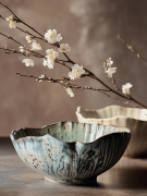 Rustykalna ceramiczna miska z melanżowym niebiesko-beżowym szkliwem stoi na ciemnym blacie. Naturalną kompozycję dopełniają kolejna miska oraz delikatne gałązki z białymi kwiatami, miękko rozmyte w tle.