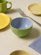 Mała, dwukolorowa żebrowana ceramiczna miseczka w kolorze miętowym z gładkim lawendowym wnętrzem stoi na beżowym stole. Obok stoją żółte talerzyki, zielony kubek, serwetka oraz widelec.