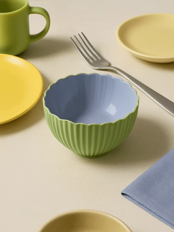 Mała, dwukolorowa żebrowana ceramiczna miseczka w kolorze miętowym z gładkim lawendowym wnętrzem stoi na beżowym stole. Obok stoją żółte talerzyki, zielony kubek, serwetka oraz widelec.