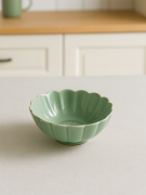 Ceramiczna, szkliwiona miseczka w kolorze szałwiowej zieleni o kształcie kwiatu stoi na jasnym blacie. Jasna aranżacja kuchenna przedstawia miękko rozmyte białe szafki i drewniany blat w tle.