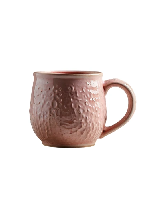 Różowy kubek ceramiczny o pękatym kształcie i zaokrąglonym uchu na jednolitym białym tle. Korpus ma mocno teksturowaną, wgłębioną powierzchnię przypominającą kratery, pokrytą błyszczącym szkliwem.