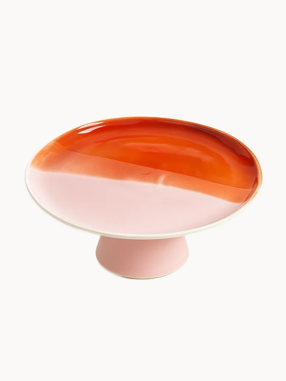Ceramiczna patera z dwukolorową powierzchnią na białym tle. Gładki blat jest w połowie pokryty błyszczącym, bursztynowo-pomarańczowym szkliwem, a w połowie matowym, pastelowym różem, osadzonym na krótkiej różowej nóżce.